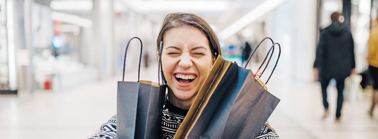 Nainen on ostoskeskuksessa ja nauraa silmät kiinni kaksi paperikassia kainaloissaan.