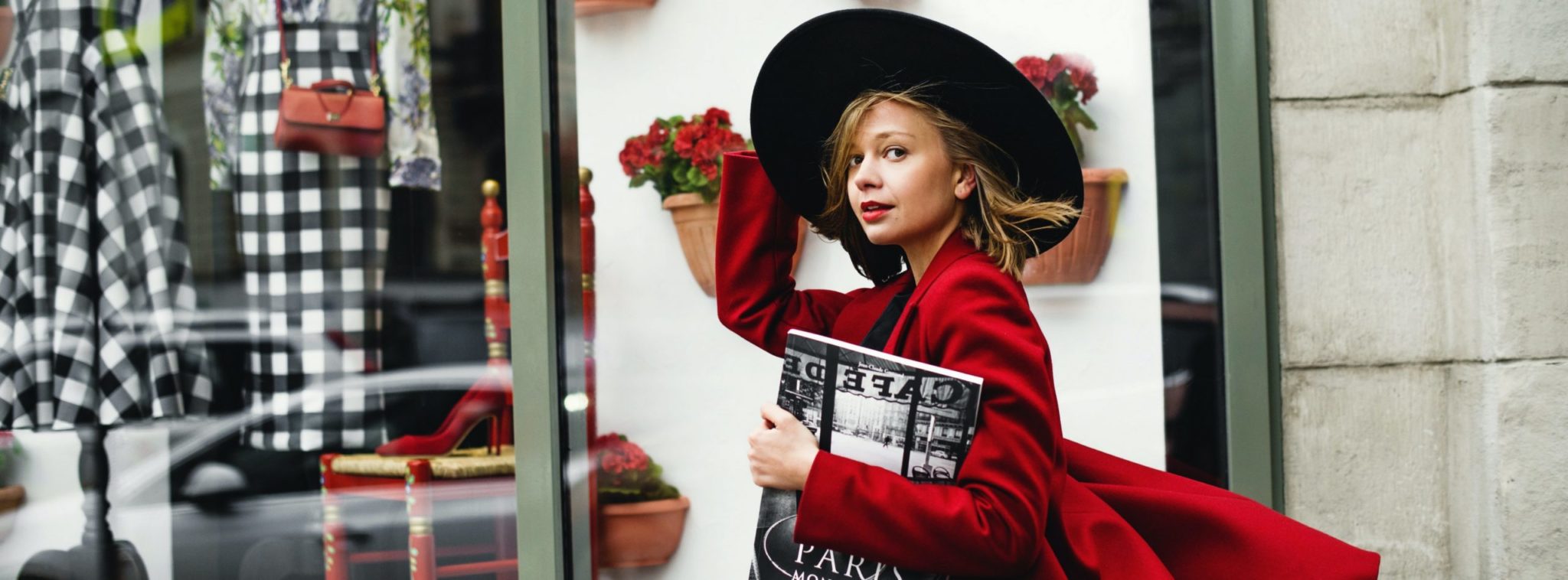 Nuori nainen kävelee vaateliikkeen näyteikkunan ohi ja katsoo kameraan punainen takki yllään ja musta lierihattu päässään, aikakauslehti kainalossa.