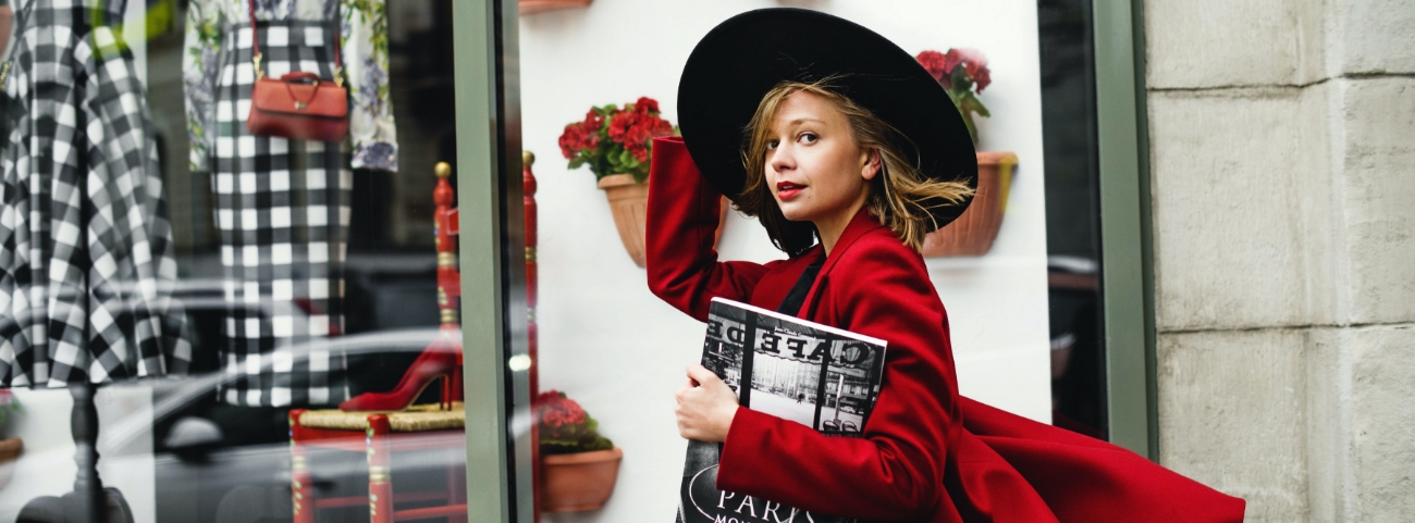 Nuori nainen kävelee vaateliikkeen näyteikkunan ohi ja katsoo kameraan punainen takki yllään ja musta lierihattu päässään, aikakauslehti kainalossa.