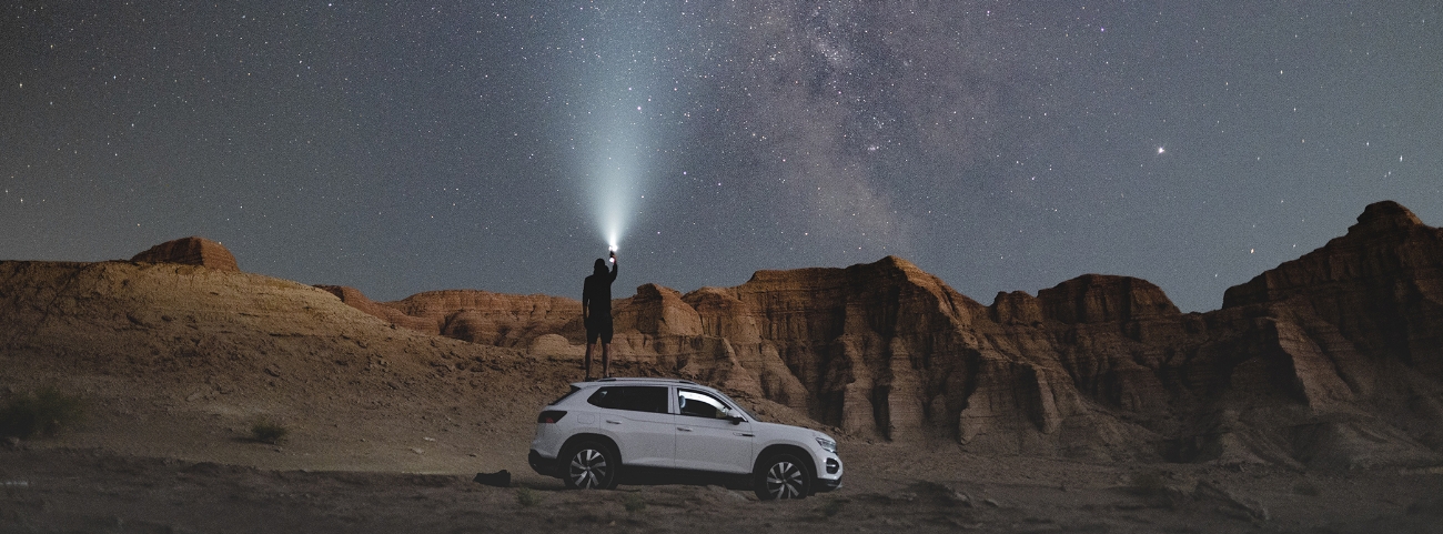 Mies on öisellä aavikolla ja seisoo valkoisen auton päällä osoittaen taskulampulla tähtitaivasta, josta erottuu linnunrata.
