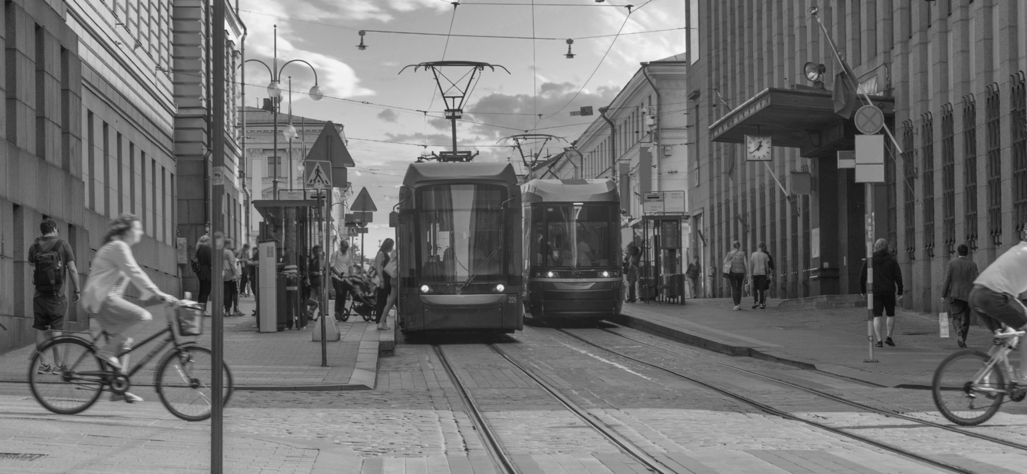 Kuva on mustavalkoinen ja siinä on katunäkymä Helsingin keskustasta, Aleksanterinkadulta. Kuvassa näkyy kaksi raitiovaunua